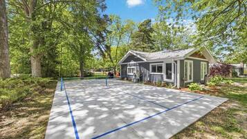 A residential property with a paved driveway, a small house with a gabled roof, and a yard with trees and shrubs.