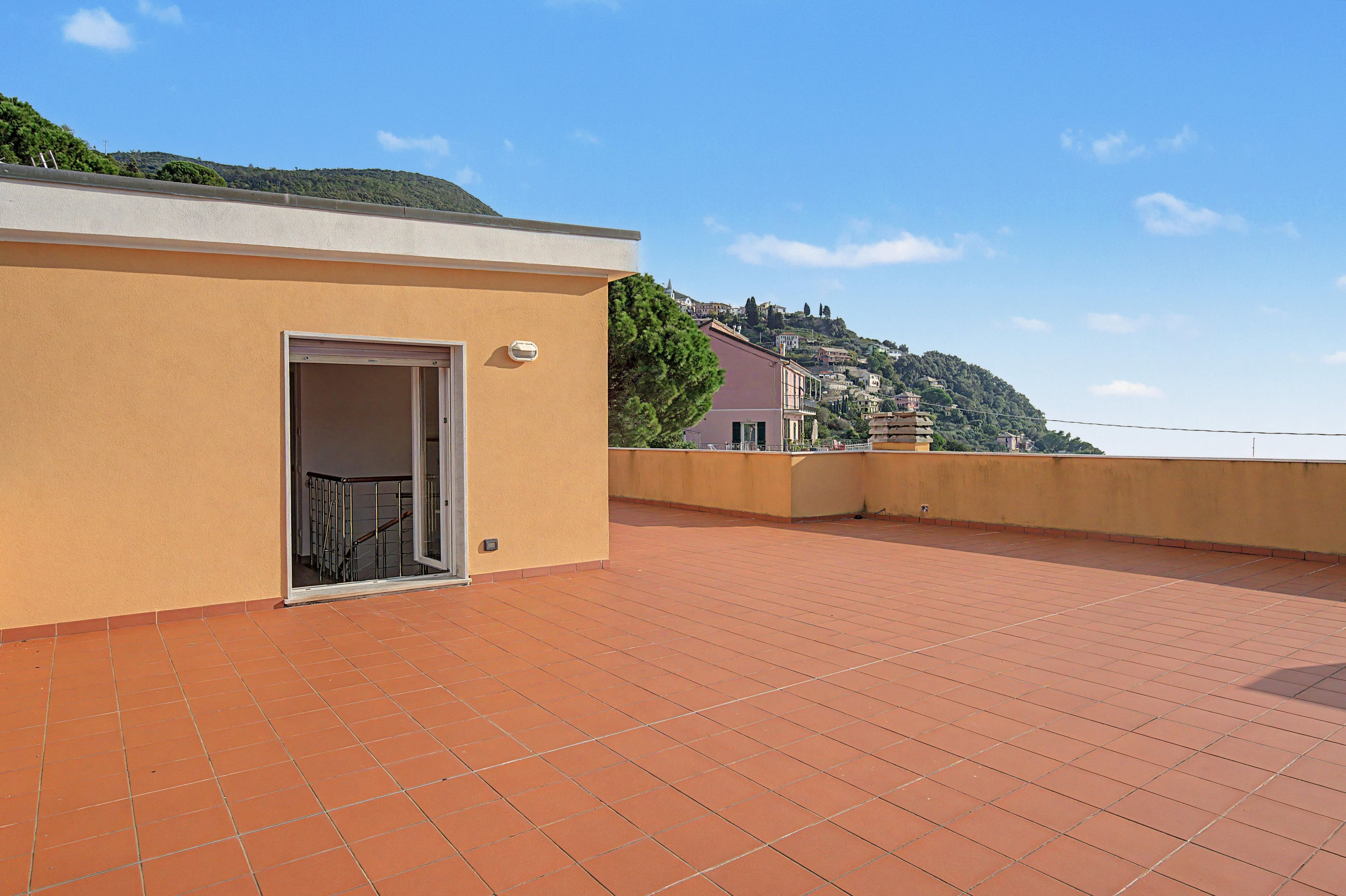 A rooftop terrace with a door leading to a balcony, a tiled floor, and a view of a hillside with buildings.