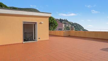 A rooftop terrace with a door leading to a balcony, a tiled floor, and a view of a hillside with buildings.
