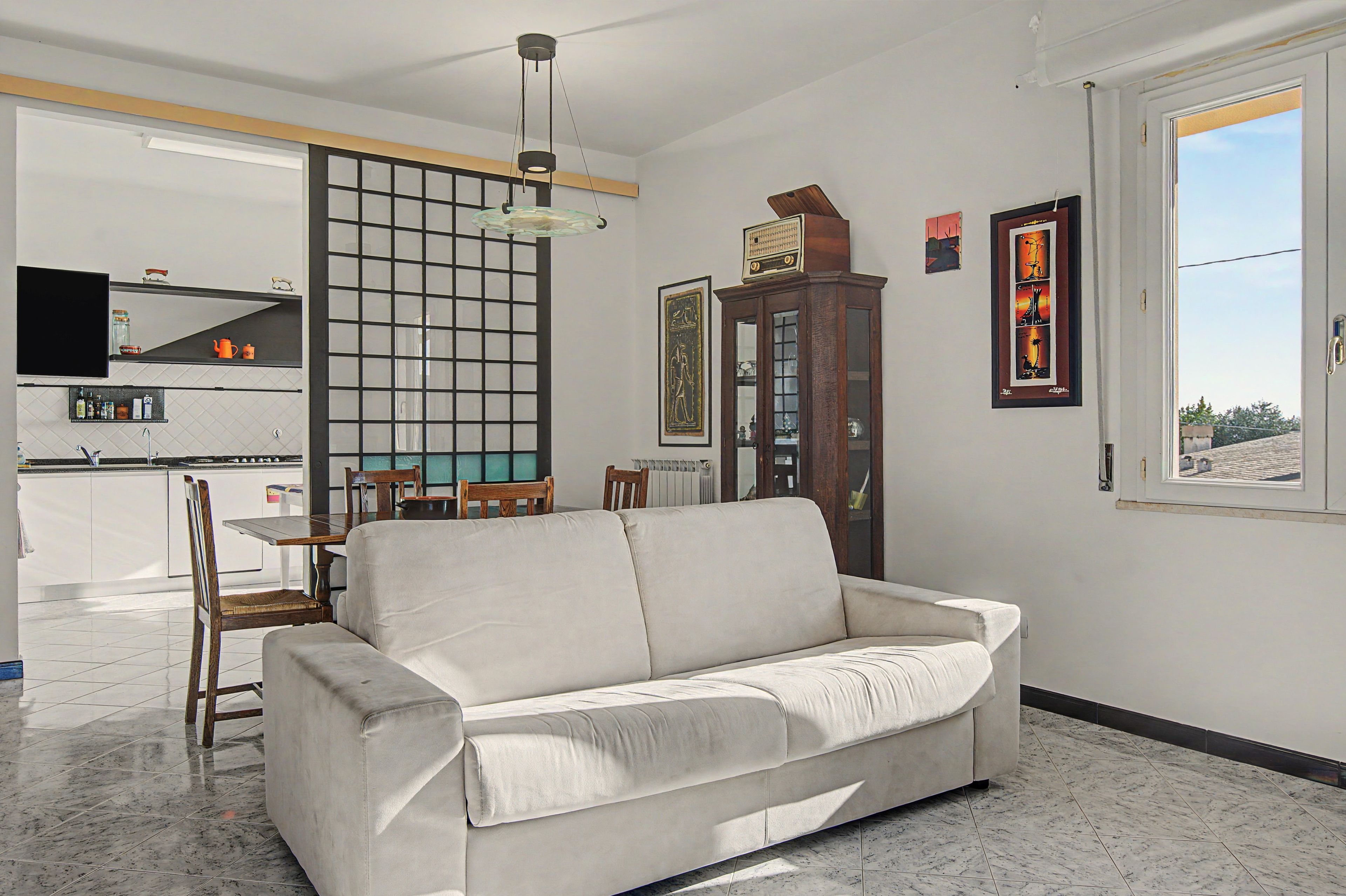 A living room with a sofa, a dining area with a table and chairs, a kitchen with a TV, and a large window.