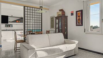 A living room with a sofa, a dining area with a table and chairs, a kitchen with a TV, and a large window.