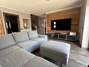 A modern living room with a sectional sofa, a flat-screen TV mounted on a wooden wall, and a staircase leading to another level. - Il Riparo – Silence and Luxury in the Mountains (Le Lou)