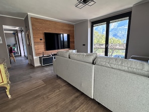 A modern living room with a large flat-screen TV mounted on a wooden wall, a grey sectional sofa, and a sliding glass door leading to a balcony with a mountain view. - Il Riparo – Silence and Luxury in the Mountains (Le Lou)