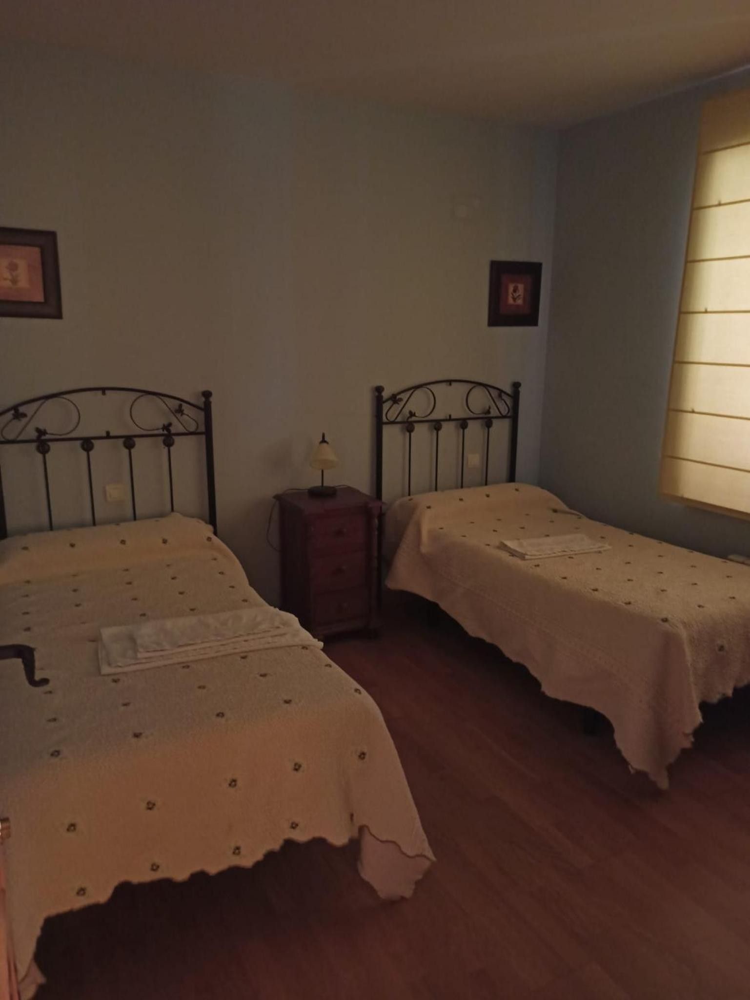 A room with two single beds, a bedside table with a lamp, and a window with blinds.