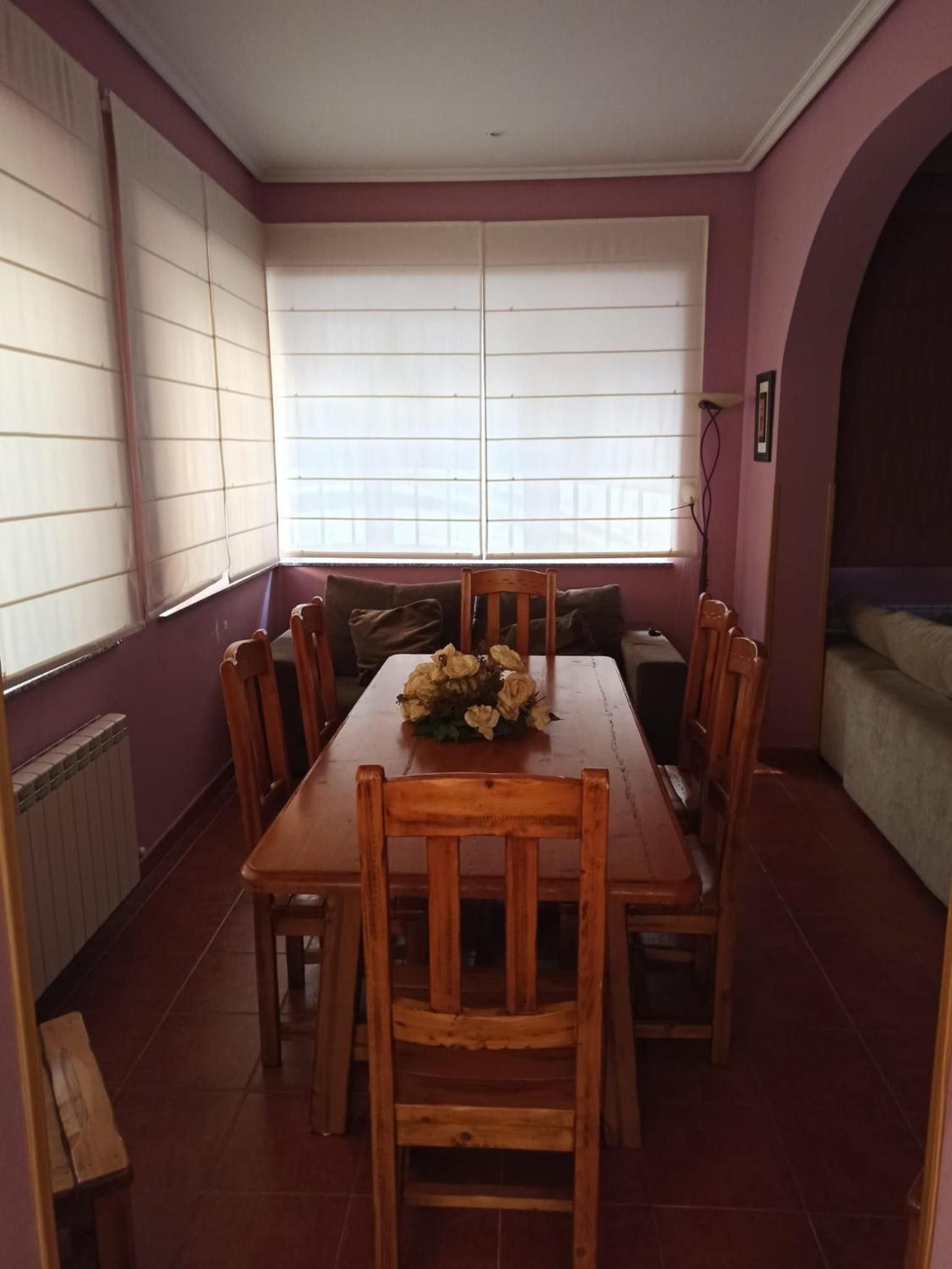 A dining area with a wooden table and chairs, a flower centerpiece, and large windows with blinds.
