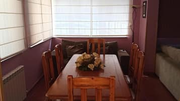 A dining area with a wooden table and chairs, a flower centerpiece, and large windows with blinds.
