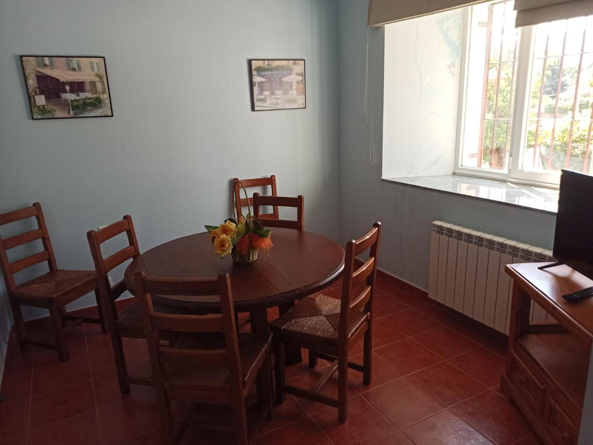 A dining area with a round table, six wooden chairs, and a vase with flowers. There are two framed pictures on the wall, a window with a view, and a radiator.