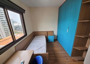 Un dormitorio pequeño con una cama individual, un escritorio, una silla, un armario y una ventana con vistas a edificios.