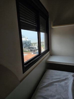 A room with a window overlooking a cityscape, a bed, and a white shelf. - 3-bedroom apartment with ocean view and 2 covered parking spaces. (Florianópolis)