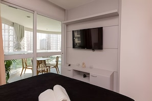 Une chambre d’hôtel avec un lit, un téléviseur fixé au mur, un balcon avec une table et des chaises, et une fenêtre avec des rideaux.