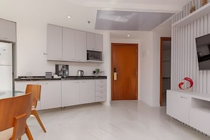A modern kitchen with white cabinets, a wooden dining table, and a TV mounted on a white wall. - Apartamento Espaçoso e Confortável (Rio de Janeiro)