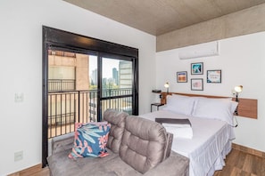 Quarto com cama, sofá, varanda com guarda-corpo e vista para a cidade.