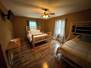 Una habitación con una cama de madera, una cama adicional, un escritorio, un ventilador de techo y un televisor montado en la pared.