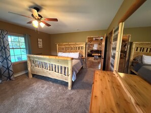 Une chambre avec un lit en bois, un ventilateur de plafond et une vue sur le salon, accessible par une porte ouverte.