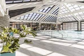 Una piscina interior espaiosa amb un sostre alt i trageters, envoltada d'àrees d'seure i plantes en testos.