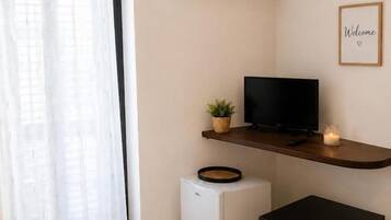 Ein Zimmer mit einem Bett, einem kleinen Fernseher auf einem Schrank, einem weißen Minikühlschrank, einem schwarzen Tisch und einem 'Welcome'-Schild an der Wand.