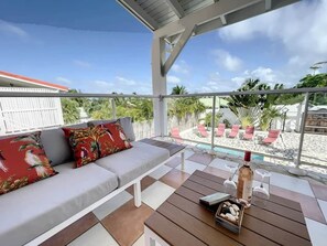 Ein überdachter Außenbereich mit einer Couch und einem Tisch, mit Blick auf einen Pool und einen Garten.