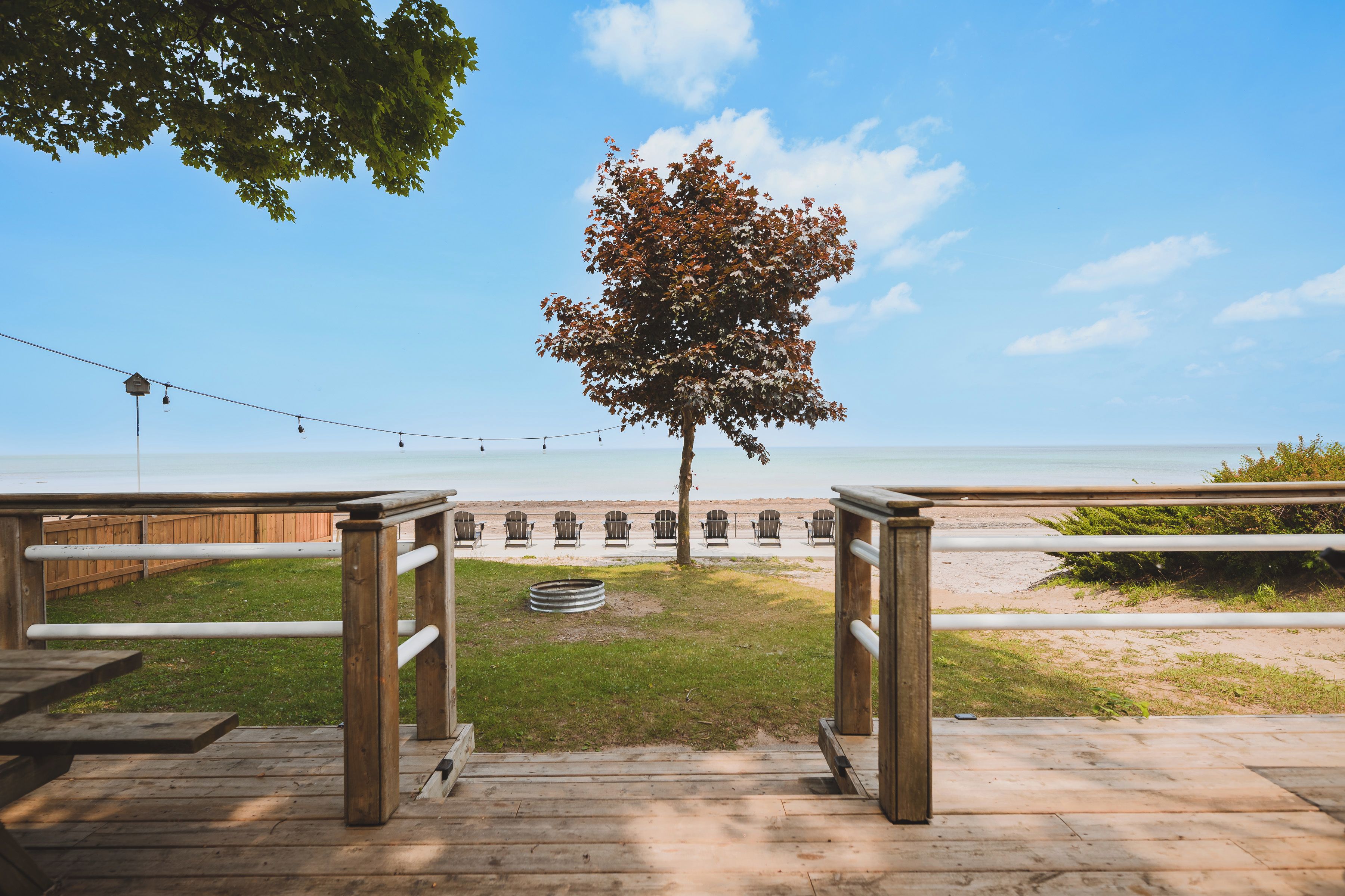 A wooden deck leading to a beach with a tree in the center, a fence, and a string of lights.