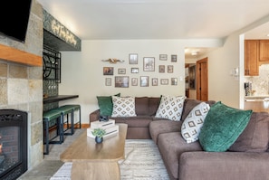 A living room with a fireplace, a brown sofa, a coffee table, and a wall decorated with framed pictures. - Modern Western Luxury in Lionshead, 2min walk to Gondola, Hot Tubs & Ski Locker (Vail)
