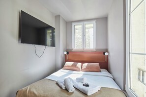 Une chambre à coucher avec un lit, un téléviseur fixé au mur, une fenêtre et une table de chevet avec une lampe.