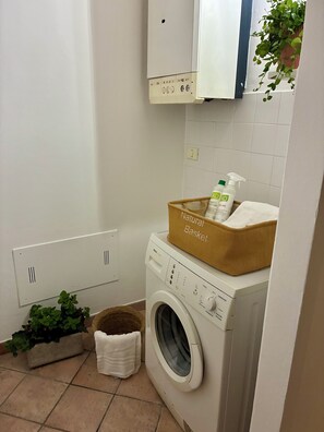 A compact laundry room with a washing machine, a basket labeled 'Natural Basket,' and a small plant. - Lovely Stay in Verona N.2 (Verona)