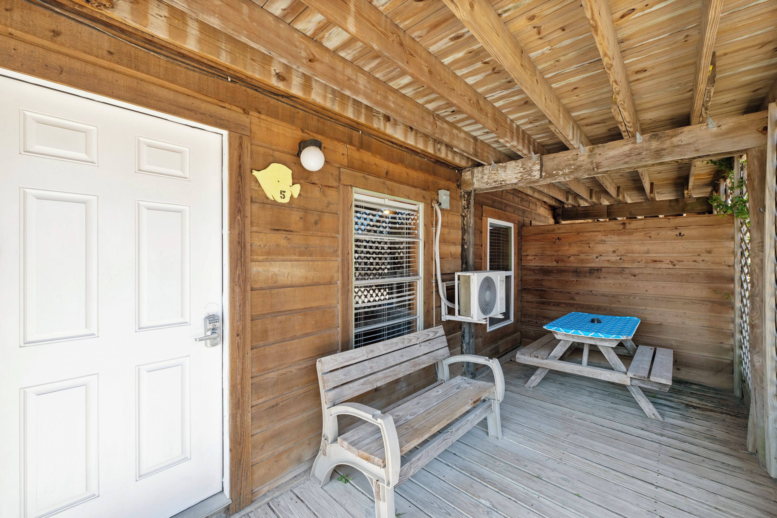 Une cabane en bois avec une porte blanche, un banc, une table recouverte d’une nappe à motifs bleus et un climatiseur fixé au mur.