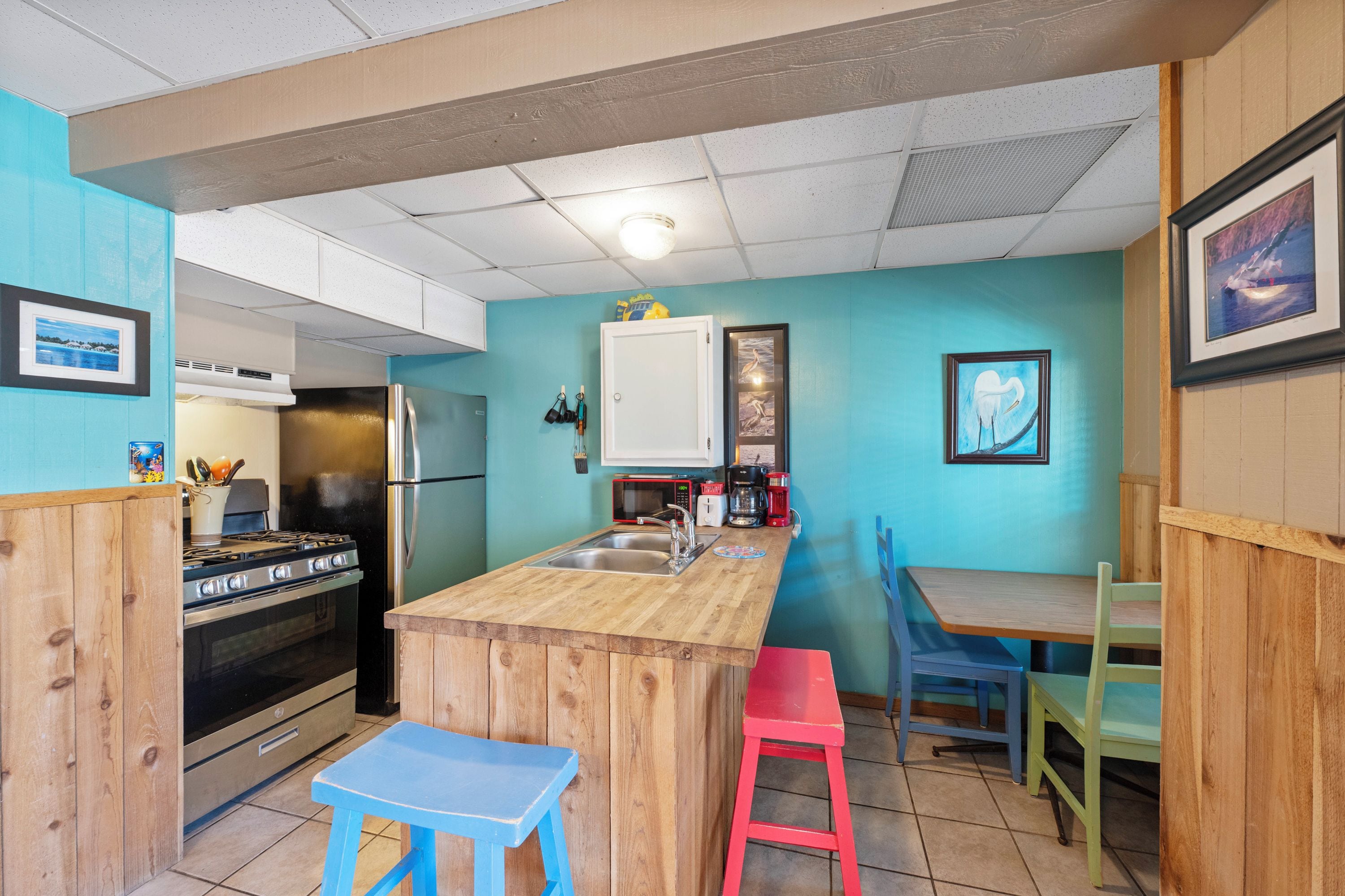 Une cuisine compacte avec un îlot central, une cuisinière, un réfrigérateur et un four à micro-ondes. On y trouve des tabourets colorés et une petite table.