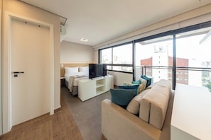 A modern hotel room with a bed, a TV, a sofa, and a large window offering a city view. - Design by Bhomy (São Paulo)