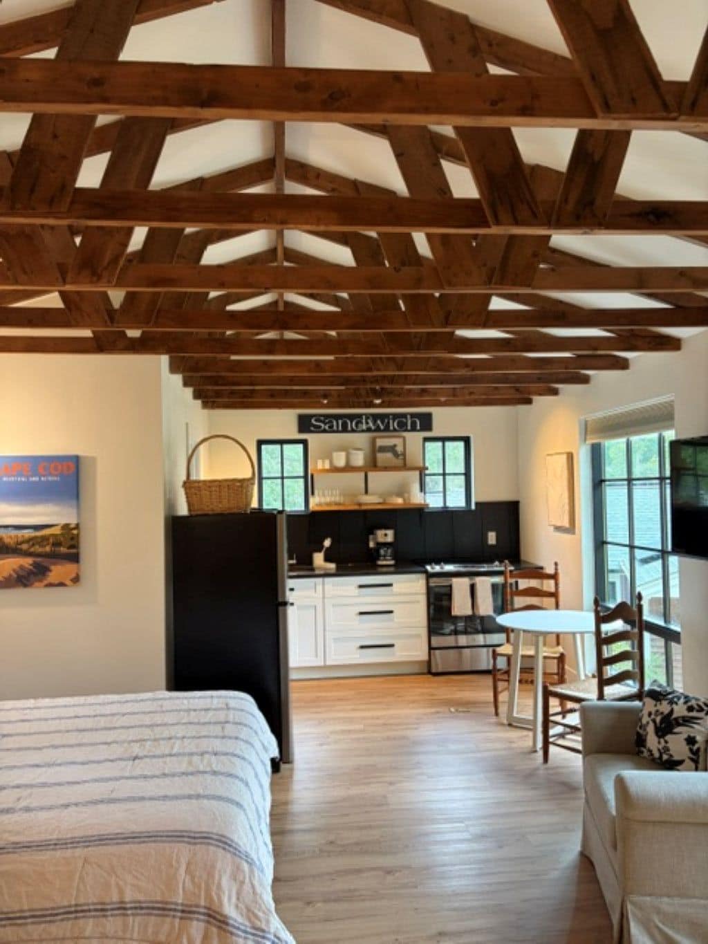 A room with a bed, a dining area with a table and chairs, a kitchen area with a sign reading 'Sandwich', and a large window providing natural light.