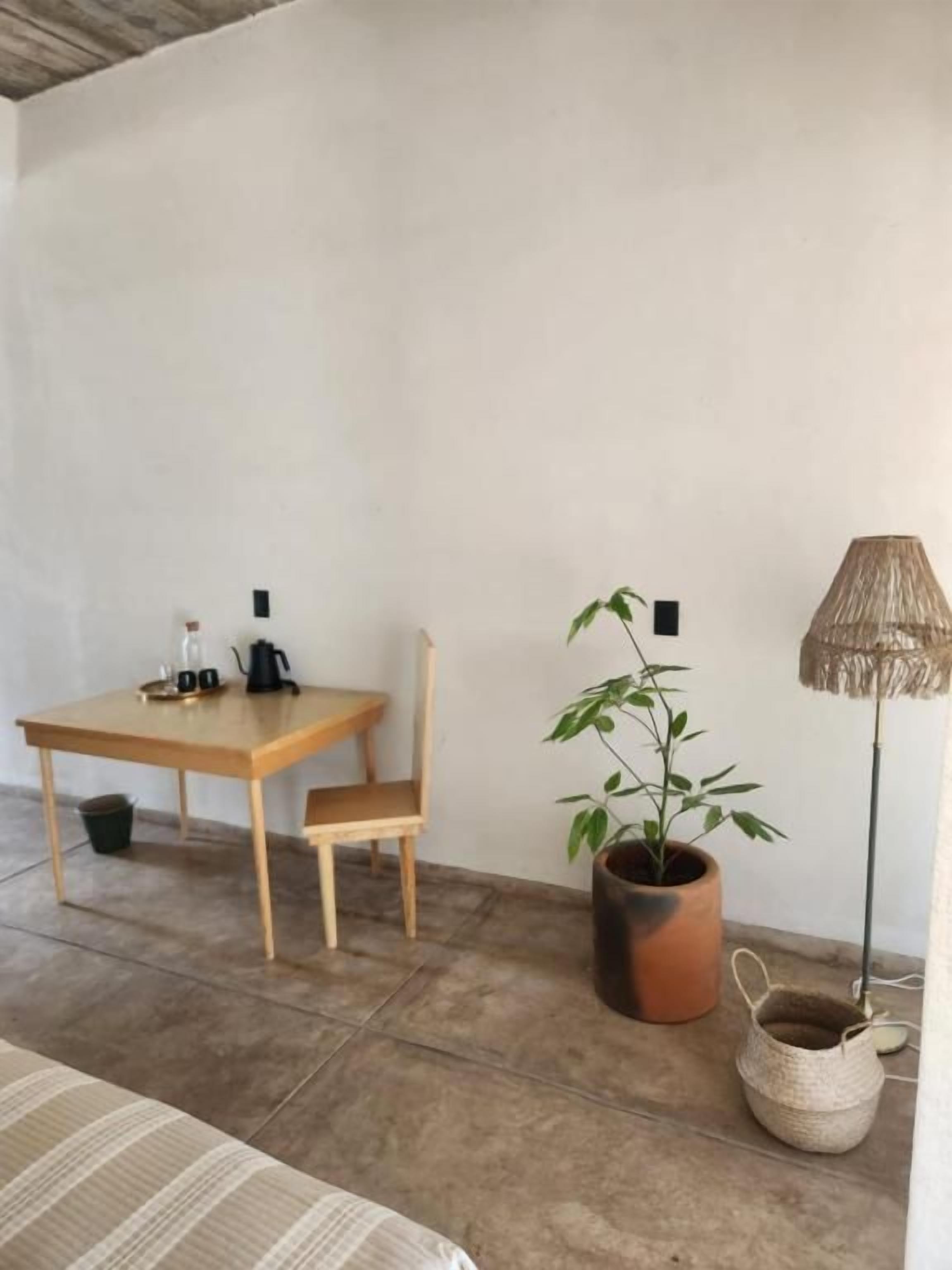 Una habitación con cama, una mesa de madera con sillas, una planta en maceta, una lámpara de pie y una cesta tejida.
