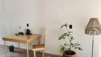Una habitación con cama, una mesa de madera con sillas, una planta en maceta, una lámpara de pie y una cesta tejida.