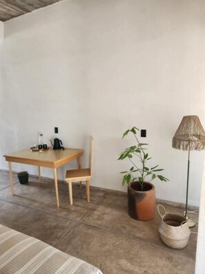 Una habitación con cama, una mesa de madera con sillas, una planta en maceta, una lámpara de pie y una cesta tejida.