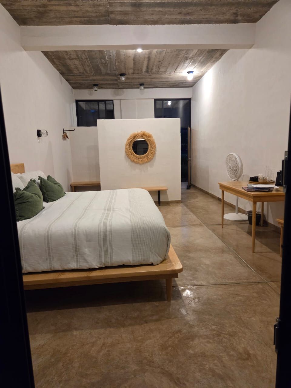 A bedroom with a bed, a wooden bench, a desk with a fan, and a circular wall decoration.
