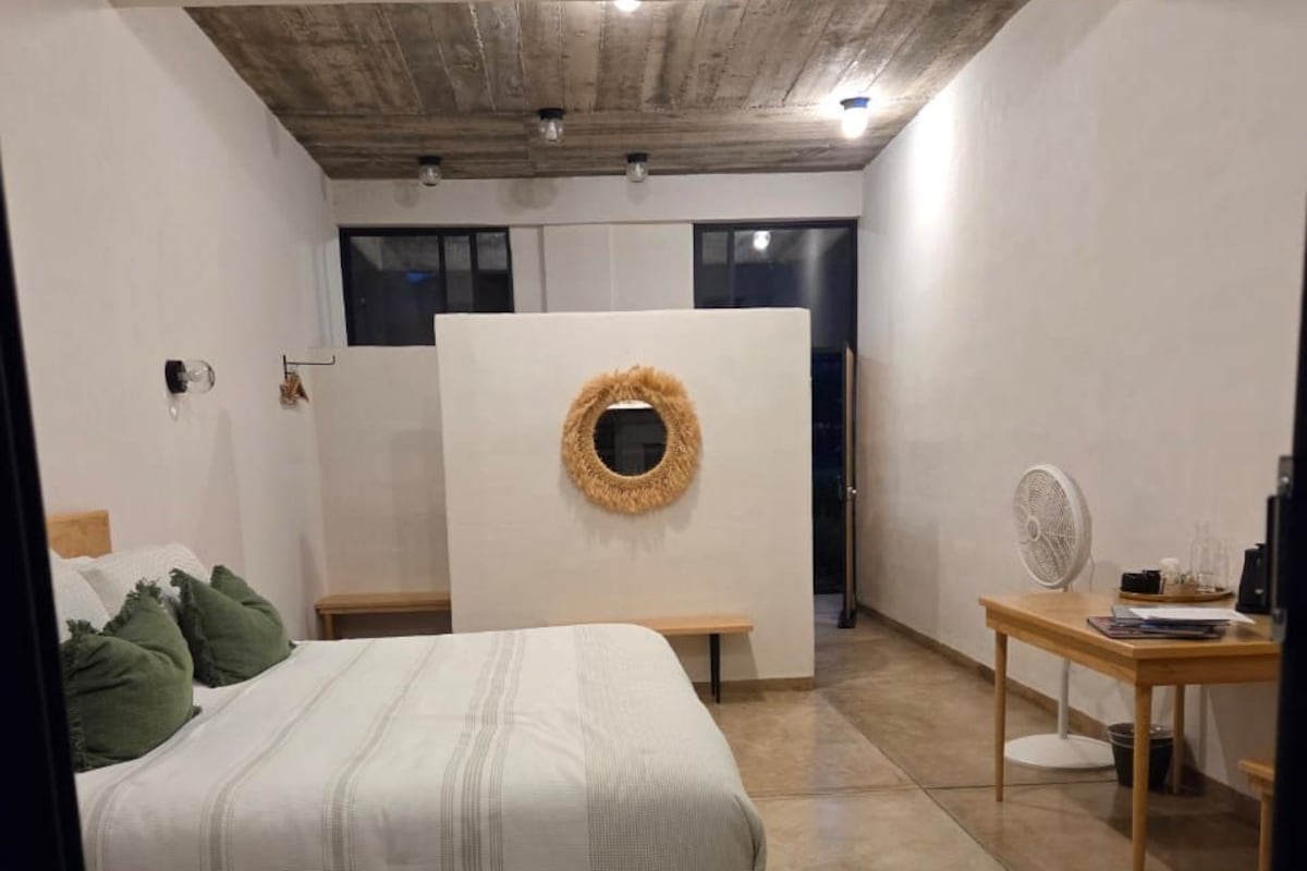 A bedroom with a bed, a wooden bench, a desk with a fan, and a circular wall decoration.