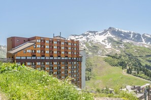 Ein mehrstöckiges Hotel mit modernem Design, das Balkone und große Fenster bietet, vor einer Kulisse aus schneebedeckten Bergen und blauem Himmel.