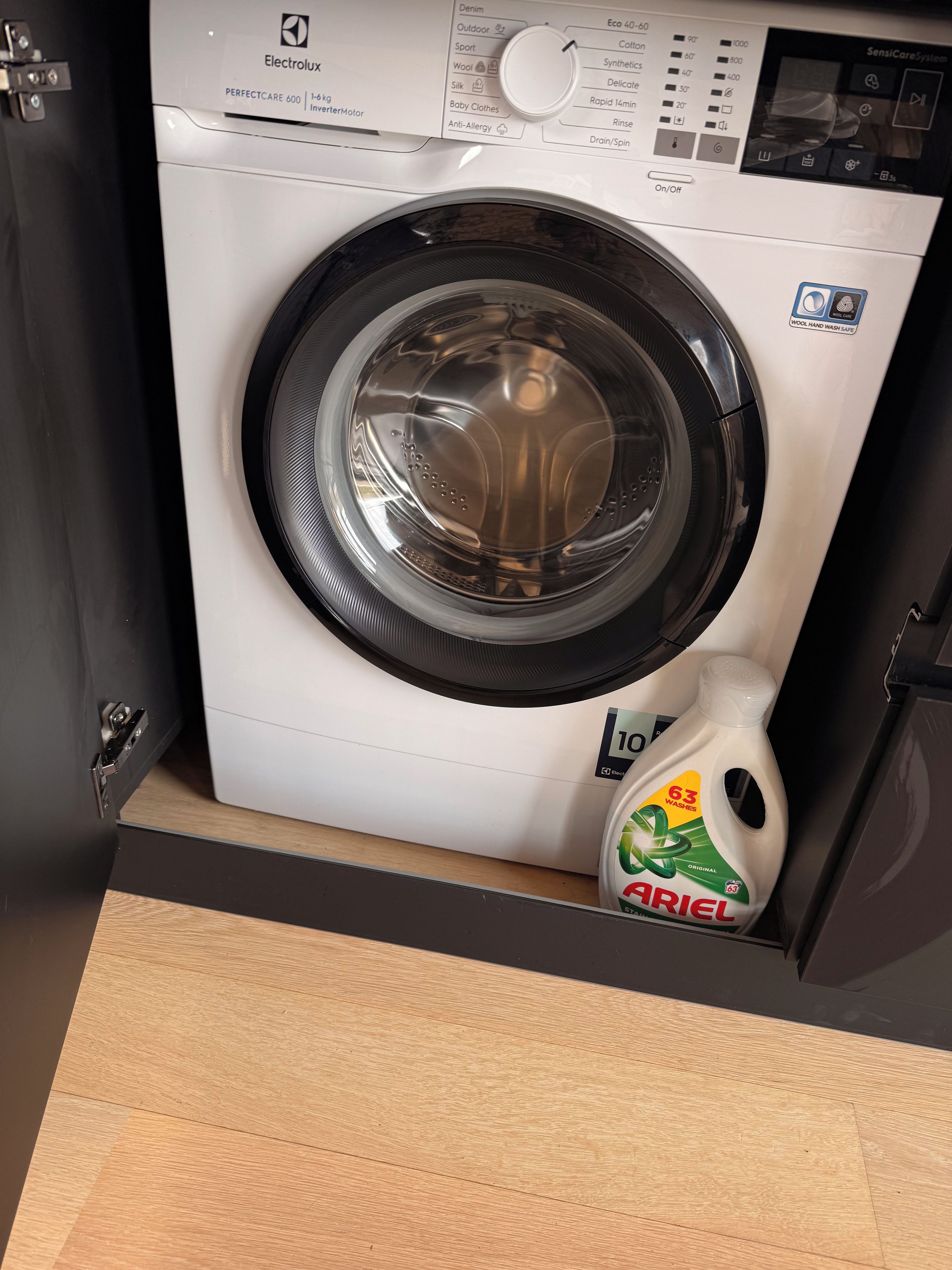 Un lave-linge à chargement frontal, doté d’un panneau de commande, d’un compartiment à lessive et posé sur un plancher en bois.
