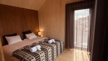 Una habitación con techo de madera, una cama grande con dos almohadas, una manta y una toalla sobre la cama. Hay una ventana con cortinas.