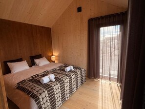 Een kamer met een houten plafond, een groot bed met twee kussens, een deken en een handdoek op het bed. Er is een raam met gordijnen.