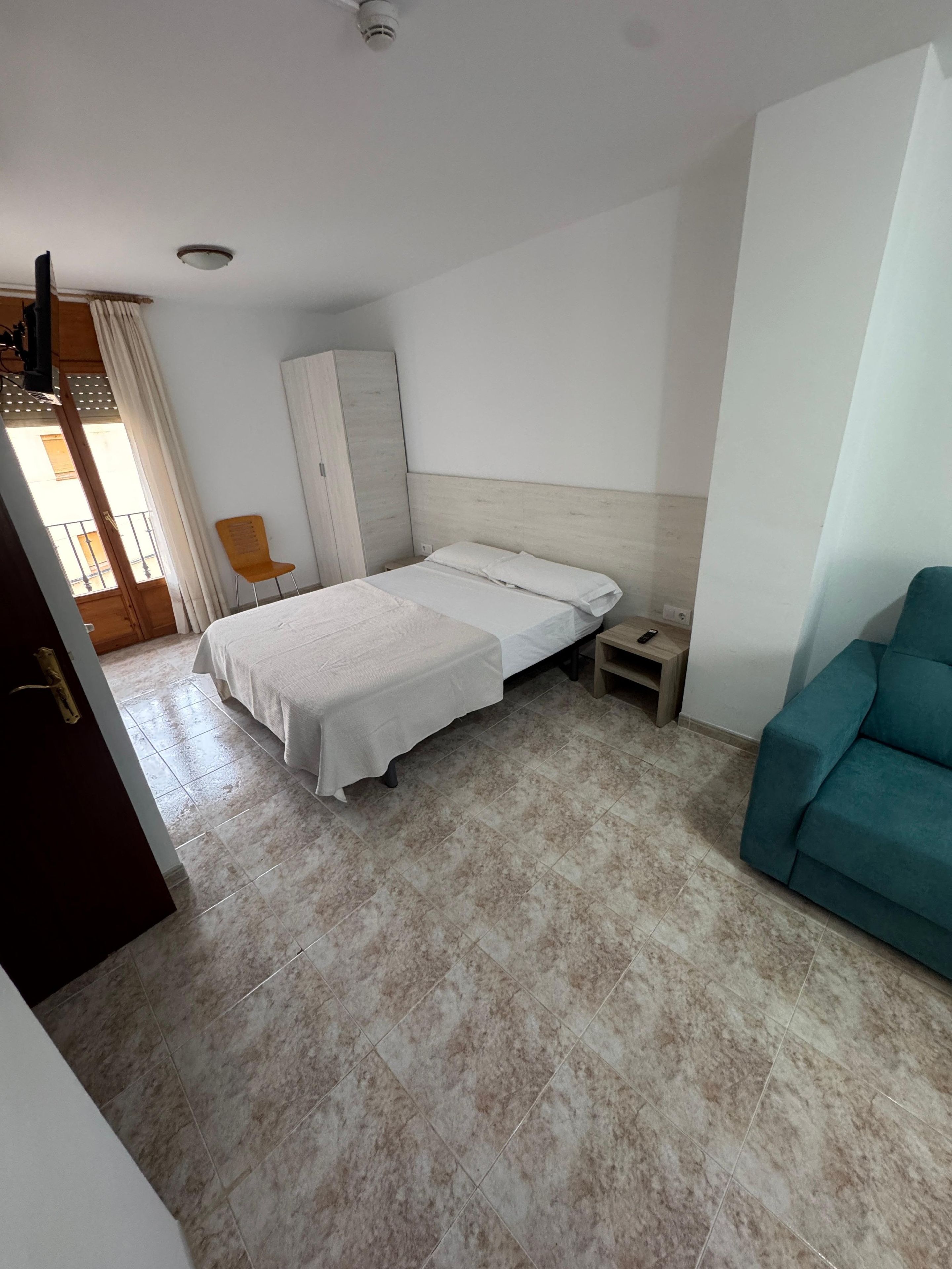 Habitación de hotel con cama, mesita de noche, sofá color turquesa, armario y ventana con cortinas.