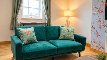 A teal sofa with floral cushions, a wooden floor, a small table, a lamp, and a floral wallpaper.