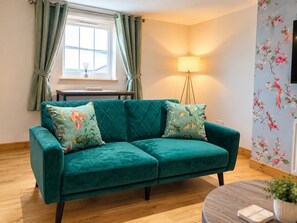 A teal sofa with floral cushions, a wooden floor, a small table, a lamp, and a floral wallpaper. - Cosy, modern top floor apartment, in the heart of Montrose (Angus)