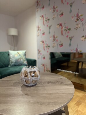 Una taula rodona de fusta amb un bol de vidre transparent ple de fruits secs, un sofà verd amb paper tapissat floral, i una làmpada al fons.