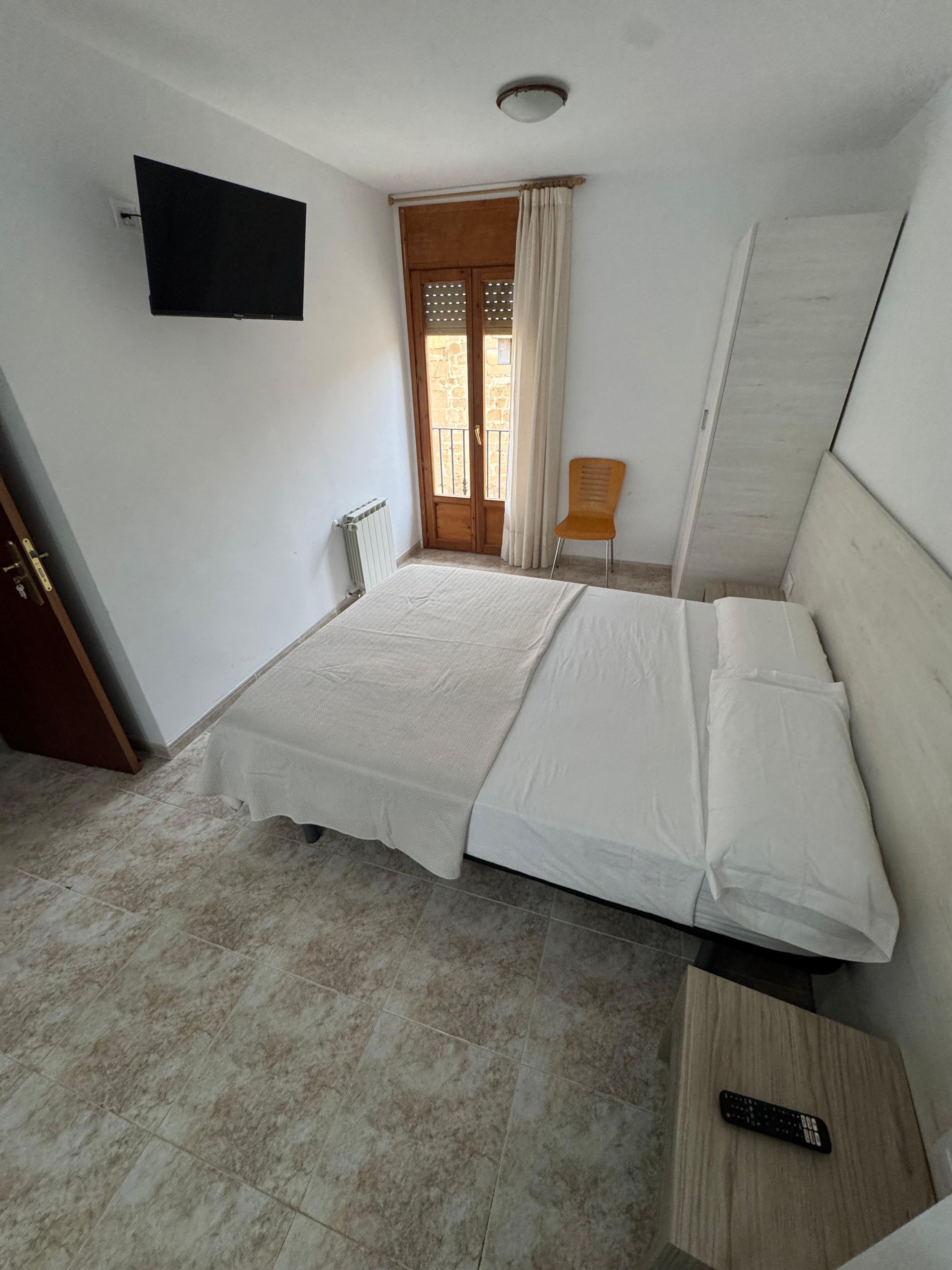 Una habitación con una cama, una silla, un televisor y un ventanal con cortinas.