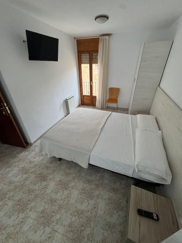Una habitación con una cama, una silla, un televisor y un ventanal con cortinas.
