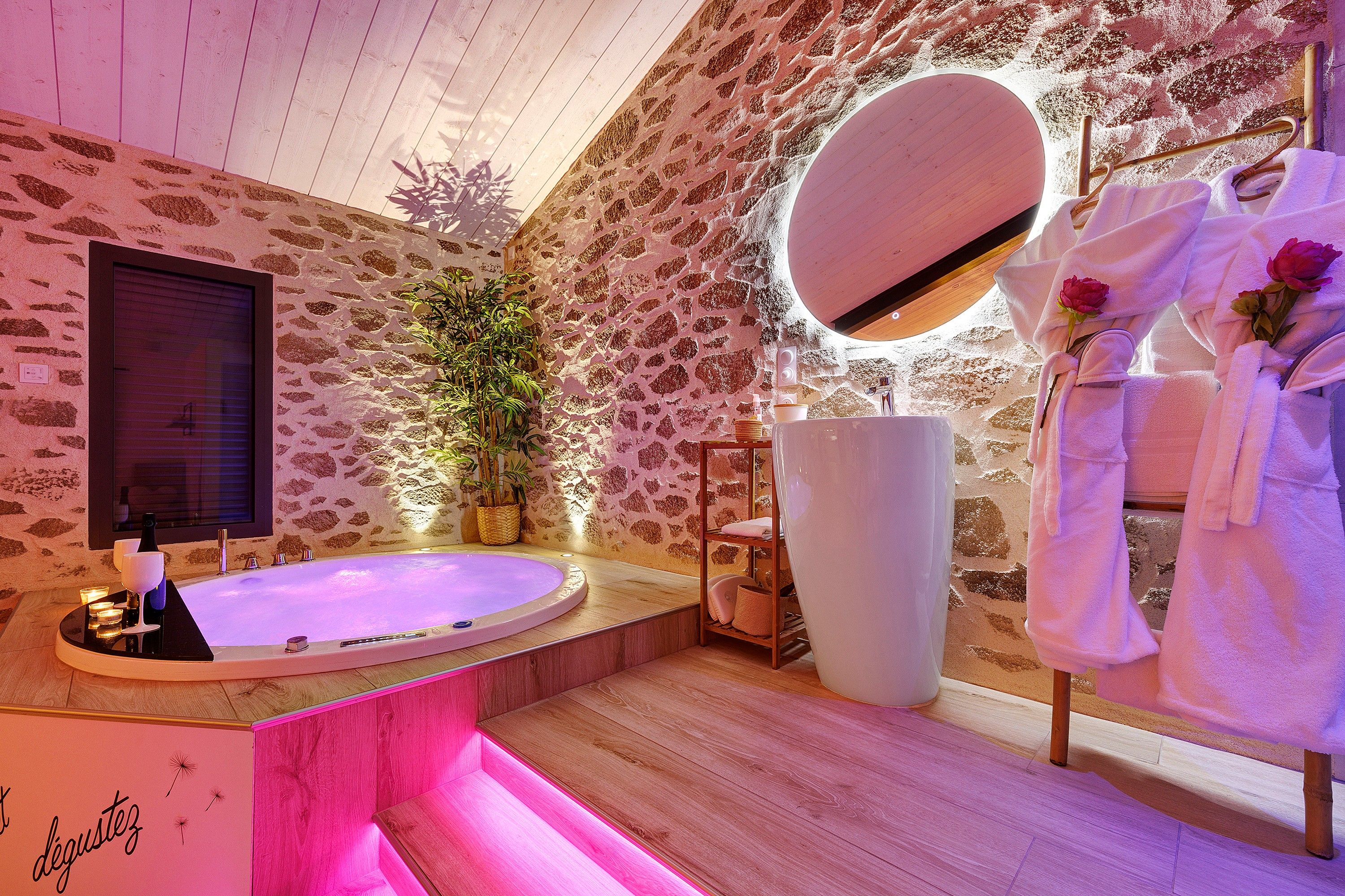 A bathroom with a stone wall, a large round mirror, a bathtub, and a wooden vanity.