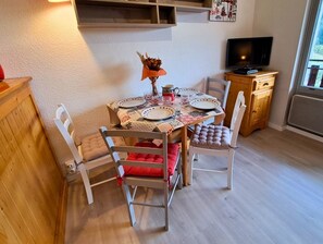 Un coin repas avec une table dressée pour quatre personnes, entourée de chaises blanches dotées de coussins rouges. Un téléviseur est fixé au mur, à droite.