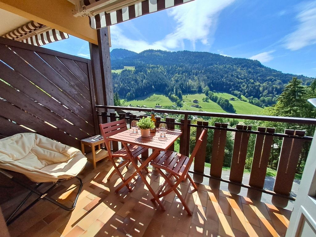 Un balcon avec une table et des chaises, un fauteuil confortable et une vue sur une vallée verdoyante et des montagnes.