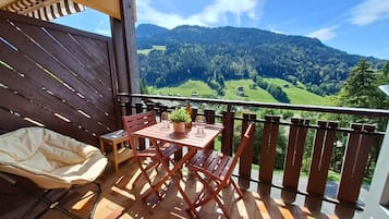 Un balcon avec une table et des chaises, un fauteuil confortable et une vue sur une vallée verdoyante et des montagnes.