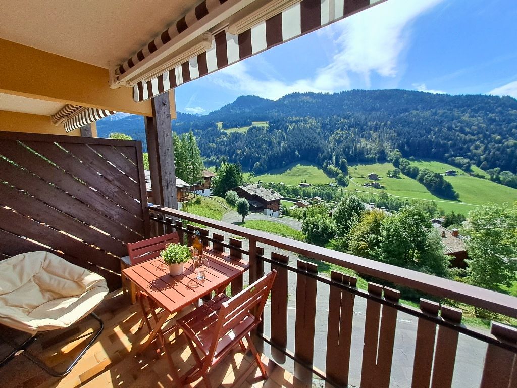 Een balkon met een houten tafel en stoelen, een bloempot en uitzicht op een groene vallei met huizen en bomen.