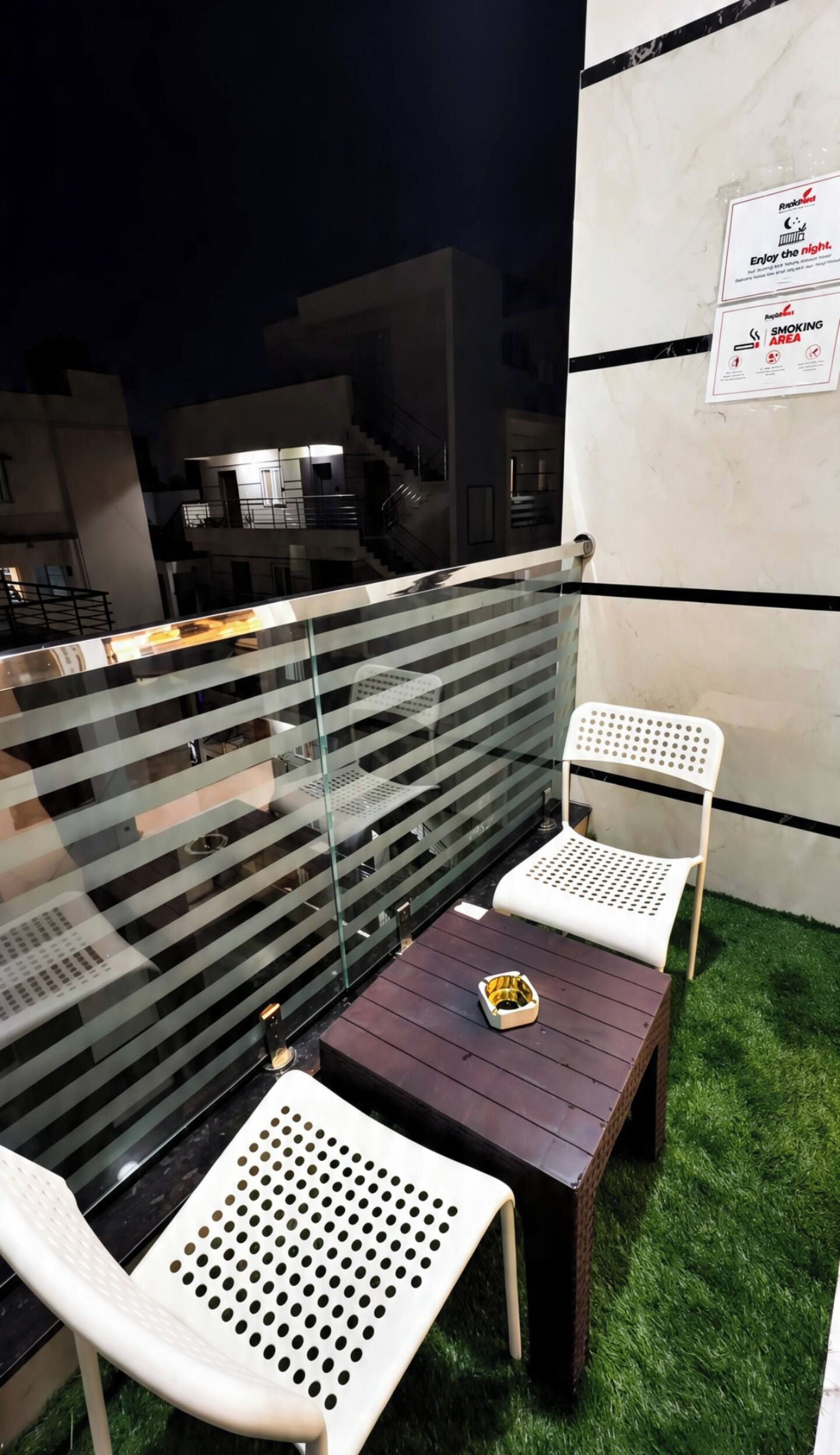 An outdoor seating area with a glass railing, two white chairs, a small table, and artificial grass.